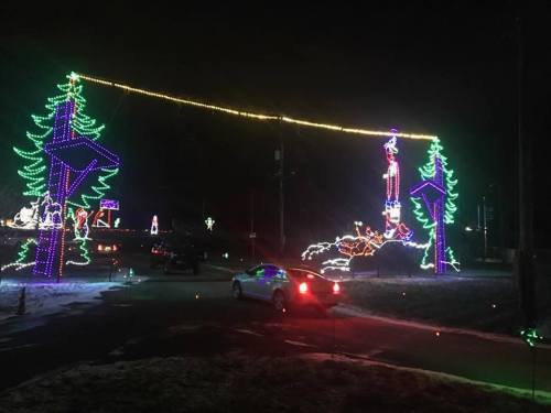 Colorful holiday lights illuminate trees and decorations in a festive nighttime scene. A car is parked nearby.