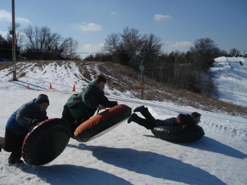 Three people sliding down a snowy hill on inflatable tubes, with trees and a clear blue sky in the background.