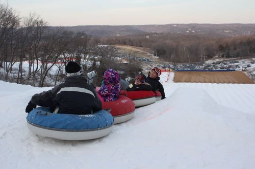 A group of people sitting on snow tubes at the top of a snowy hill, ready to slide down. Scenic winter landscape in the background.