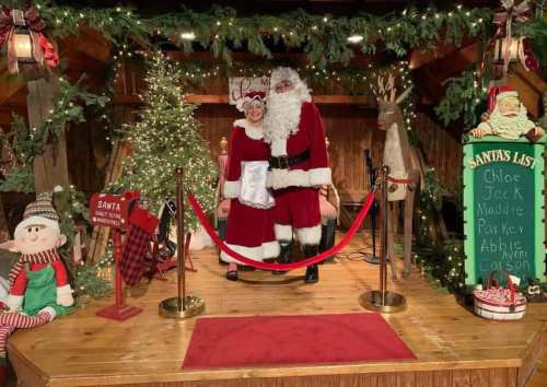 A festive scene with Santa and a woman in a red dress, surrounded by Christmas decorations and a tree.