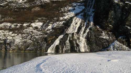 A snowy landscape featuring a cascading waterfall flowing down a rocky cliff into a calm body of water.