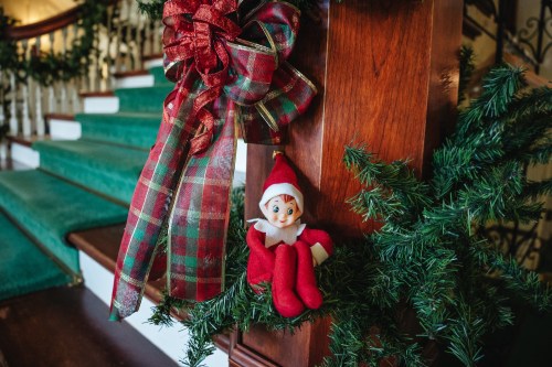 A festive staircase decorated with garlands and a cheerful elf doll sitting on the railing.