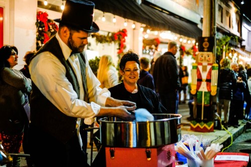 A man in a top hat prepares cotton candy at a festive outdoor event, while a woman watches nearby. Holiday decorations are visible.
