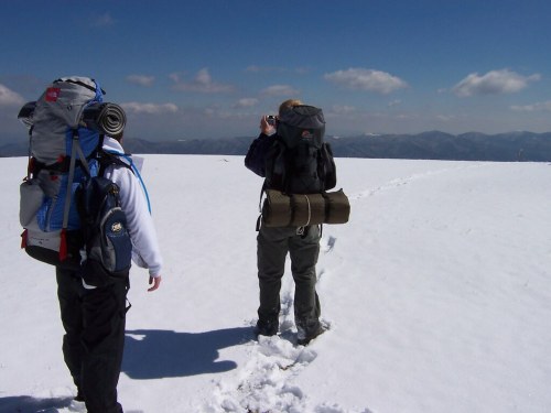 Two hikers with backpacks walking on a snowy landscape, one taking a photo against a backdrop of mountains and blue sky.