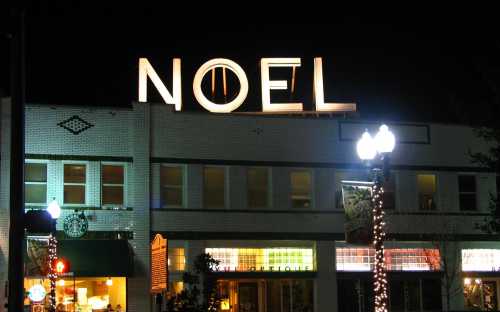 A building at night with a large illuminated "NOEL" sign and festive lights, featuring a Starbucks below.