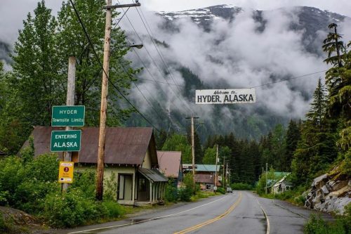 A quiet road leads into Hyder, Alaska, with misty mountains and a sign welcoming visitors to the town.