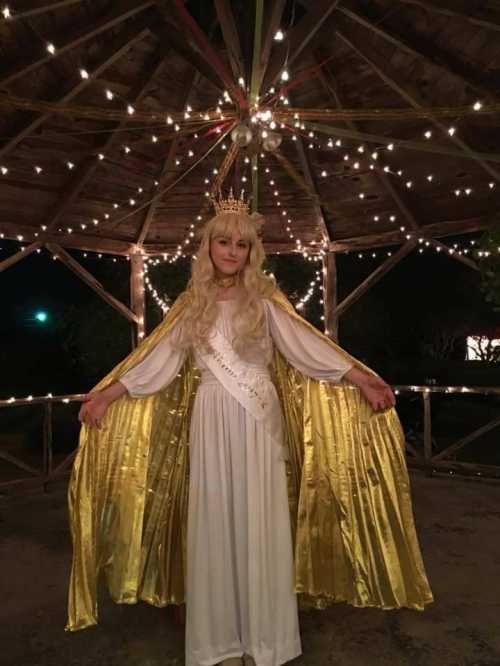 A person in a white gown and golden cape stands under a gazebo adorned with fairy lights, wearing a crown.