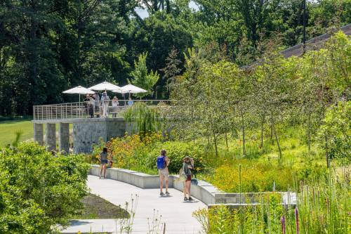 A scenic park with visitors walking along a path, lush greenery, and a shaded seating area with umbrellas.