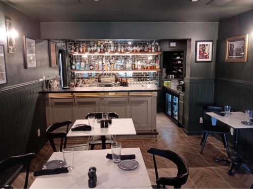 A cozy restaurant interior featuring a bar, tables with black chairs, and a well-stocked shelf of drinks.