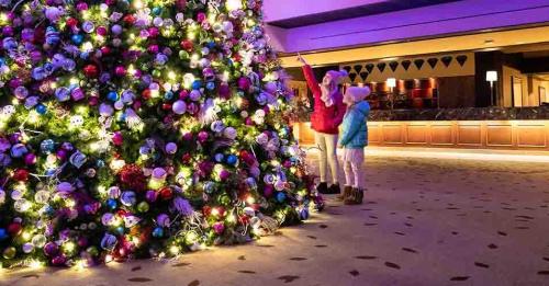 A child and an adult admire a brightly lit Christmas tree decorated with colorful ornaments in a festive setting.