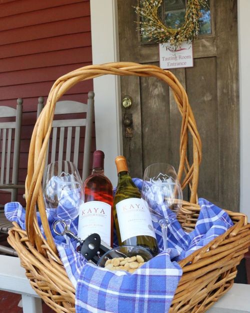 A wicker basket with two bottles of wine, glasses, and snacks, set on a porch with a welcoming entrance sign.