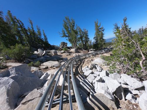 A winding roller coaster track curves through rocky terrain and trees under a clear blue sky.