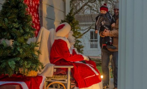 A child in winter clothing is held by a man while approaching Santa Claus on a porch decorated for Christmas.