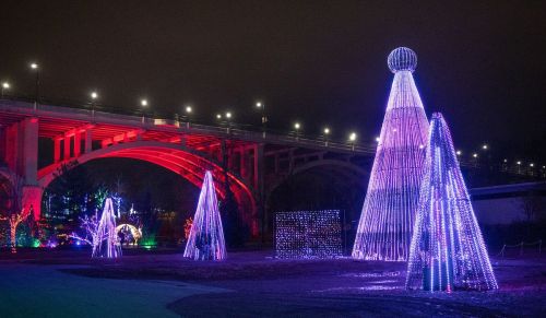 Colorful illuminated trees and a large light structure under a bridge at night, creating a festive atmosphere.