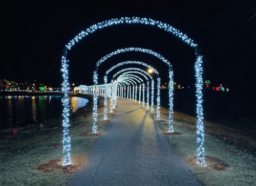 A pathway lined with glowing arches of white lights, leading towards a festive, illuminated waterfront at night.