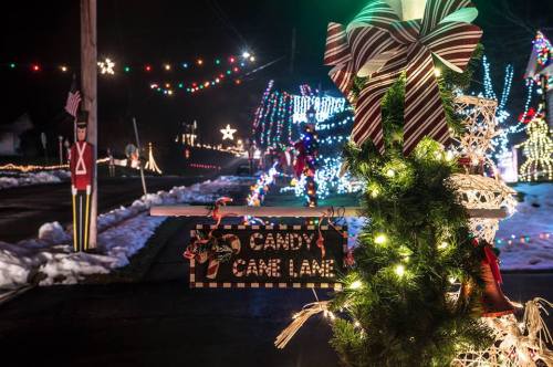 A festive street sign for "Candy Cane Lane" adorned with holiday lights and decorations in a snowy nighttime setting.