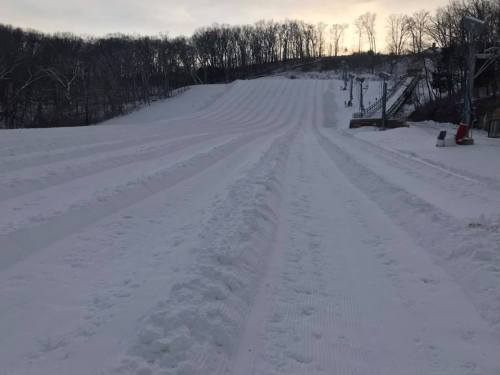 A snowy hill with groomed tracks, surrounded by trees, under a cloudy sky at dusk.