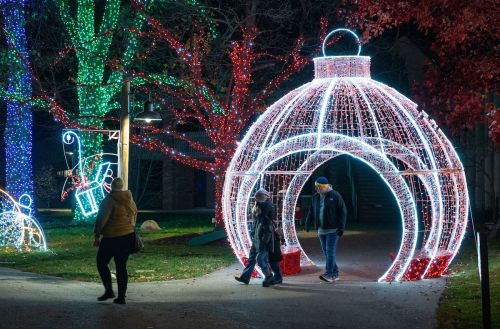 A festive scene with colorful holiday lights, featuring a large illuminated ornament and people walking through the display.