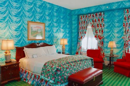 Brightly decorated hotel room with blue draped wallpaper, floral bedding, red chairs, and lamps. Cozy and vibrant atmosphere.