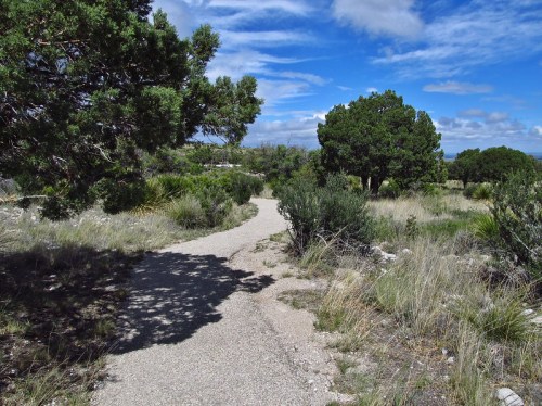 A winding gravel path through a grassy area with shrubs and trees under a blue sky with scattered clouds.