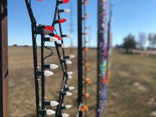 Colorful holiday lights strung along a fence, with a clear blue sky and grassy field in the background.