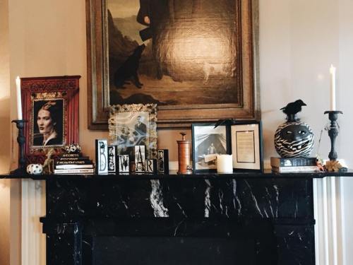 A dark marble mantel decorated with candles, framed photos, books, and a crow figurine, set against a vintage painting.