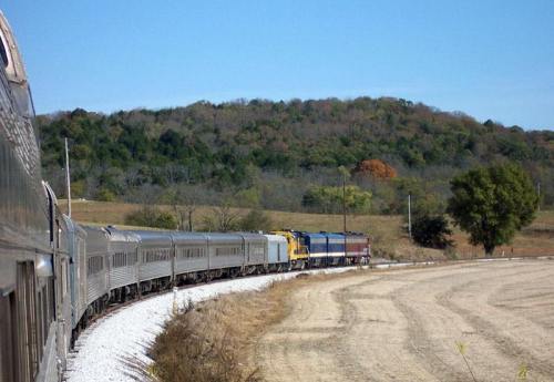 A train curves along a track with rolling hills and trees in the background under a clear blue sky.