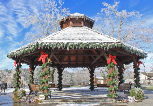A snow-covered gazebo decorated with greenery and red bows, surrounded by a winter landscape.