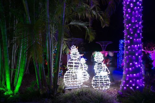 Three illuminated snowmen made of lights stand among colorful holiday decorations and palm trees at night.