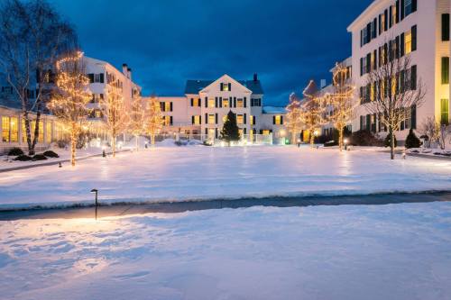 A snowy courtyard at dusk, with buildings adorned in warm lights and trees wrapped in glowing decorations.