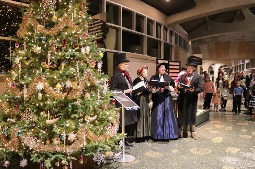 A festive scene with a decorated Christmas tree and carolers in Victorian attire singing in a holiday setting.