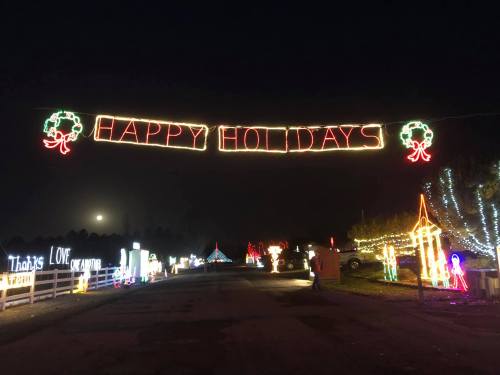 A festive display of holiday lights with a "HAPPY HOLIDAYS" sign hanging above a decorated street at night.