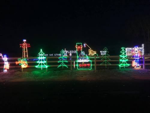 Colorful holiday lights display featuring trees, a cabin, and festive signs against a dark night sky.