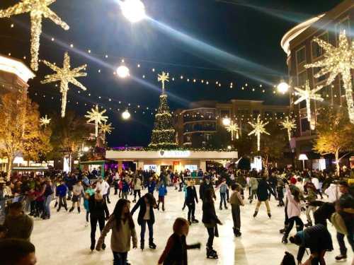 A bustling outdoor ice skating rink decorated with festive lights and a Christmas tree, filled with skaters at night.