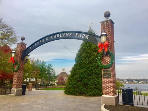 Archway entrance to Riverside Gardens Park, decorated with wreaths and bows, overlooking a calm waterway.