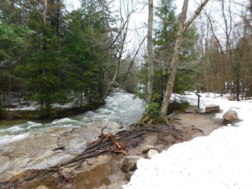 A snowy landscape with a flowing river surrounded by trees and rocky banks.