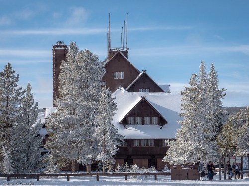 A large, rustic lodge surrounded by snow-covered trees under a clear blue sky. People are gathered in the foreground.