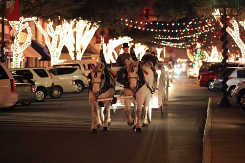 A horse-drawn carriage travels down a festive street adorned with colorful holiday lights and decorations.