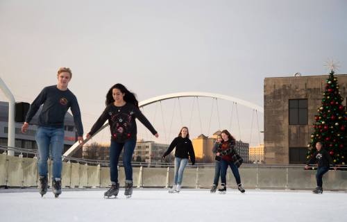 A group of five young people ice skating outdoors, with a decorated Christmas tree in the background.