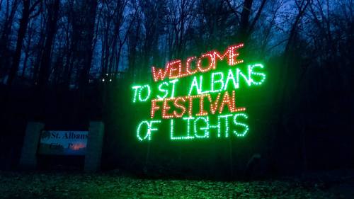 Brightly lit sign reading "Welcome to St. Albans Festival of Lights" against a dark, wooded background.