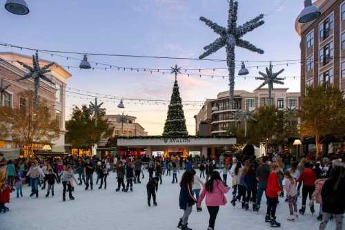 A bustling outdoor ice skating rink surrounded by festive decorations and a large Christmas tree at sunset.