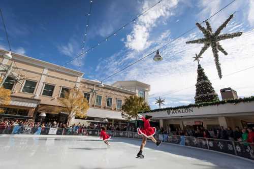 Two skaters in red dresses perform on an ice rink, with festive decorations and a large tree in the background.