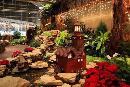 A beautifully decorated indoor garden with a small model house, rocks, plants, and festive lights.