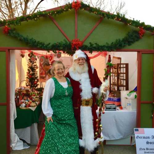 A woman in a green dress and a man in a Santa costume pose in front of a festive holiday booth decorated with greenery.