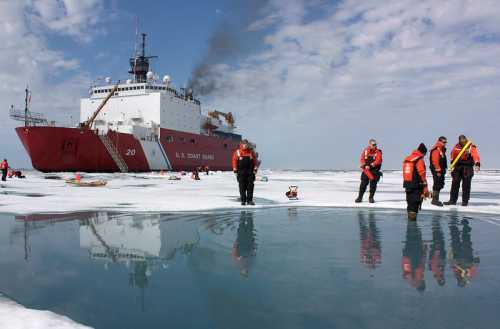 U.S. Coast Guard ship in icy waters with crew members in orange jackets examining the ice.