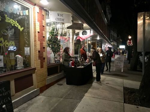 A nighttime street scene with a wine and cheese stand outside a shop, people browsing and enjoying the festive atmosphere.