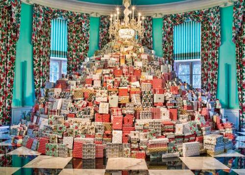 A large pyramid of colorful wrapped gifts fills a room with festive decorations and a chandelier overhead.