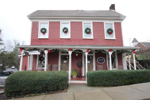 A charming red house decorated with wreaths and holiday accents, featuring a porch and green hedges.
