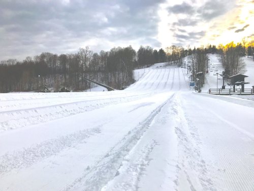 A snowy landscape with groomed slopes, trees in the background, and a cloudy sky at sunset.