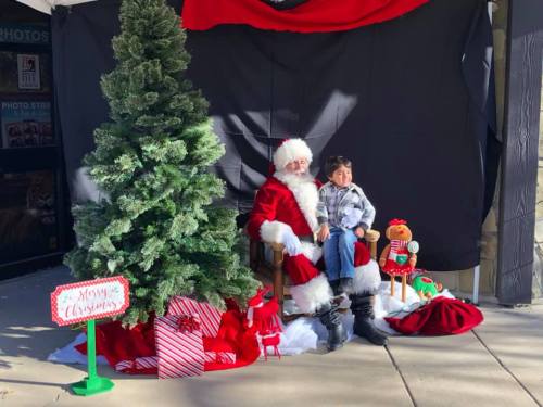 A festive scene with Santa Claus sitting next to a child, surrounded by a Christmas tree, gifts, and holiday decorations.